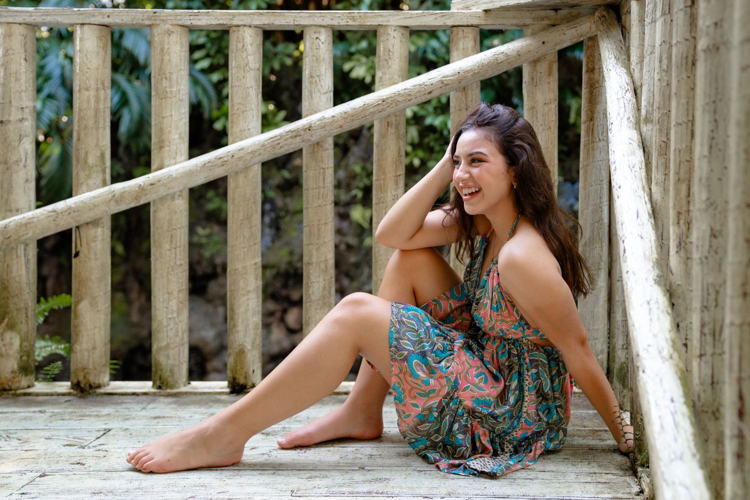 Smiling young woman in floral sundress sitting barefoot on wooden deck surrounded by lush greenery.