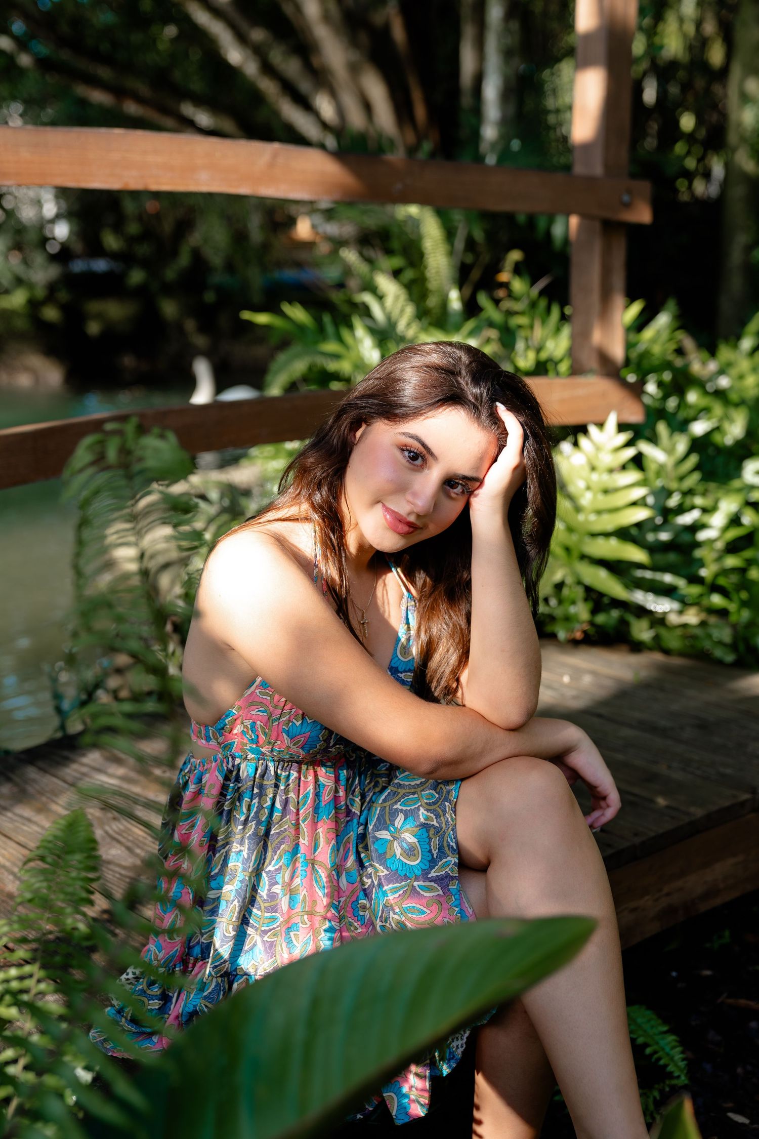 Young woman in colorful floral dress sitting on wooden deck surrounded by lush tropical garden greenery.