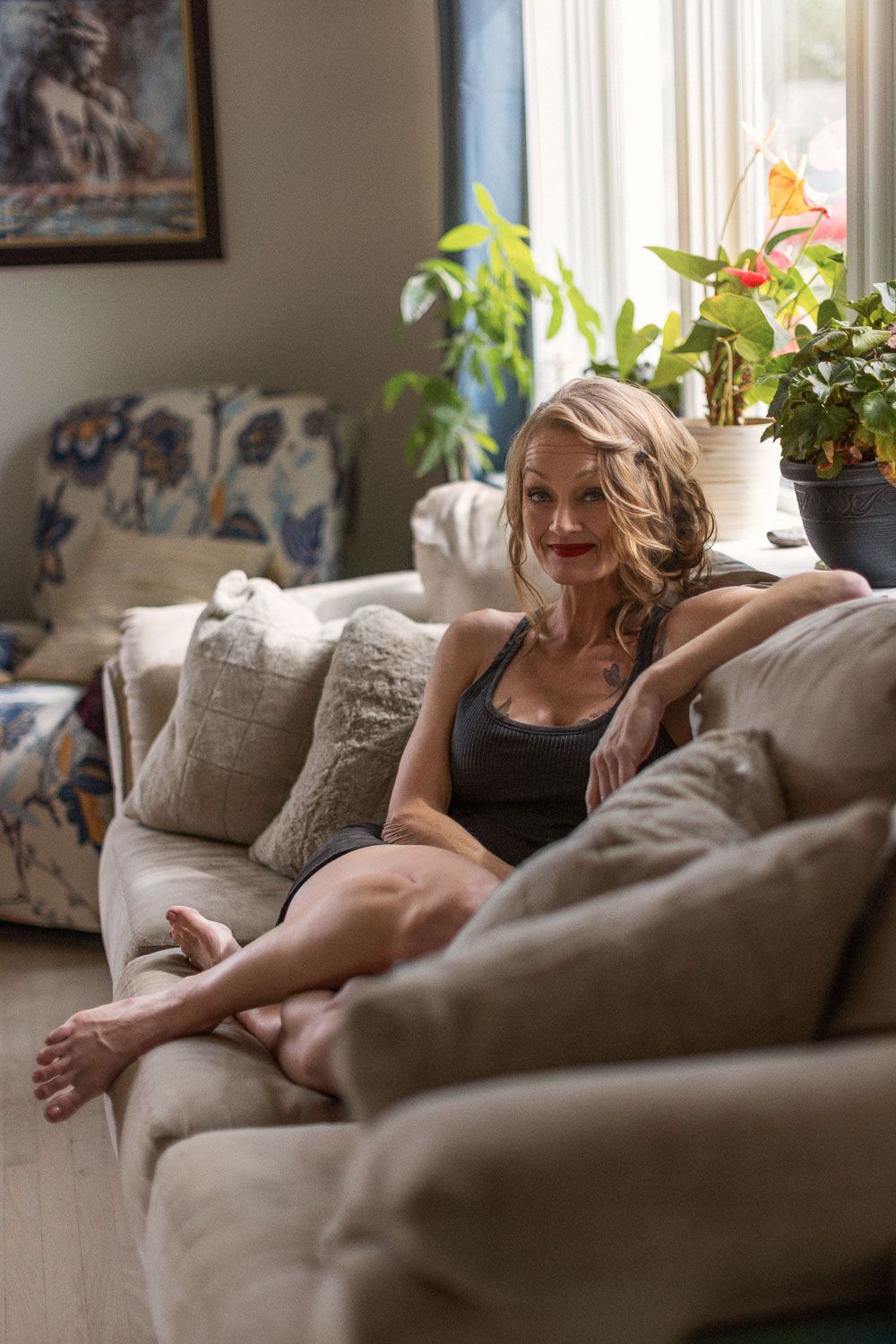 Blonde woman in gray tank top relaxing on beige sofa with pillows, plants and artwork in bright living room.