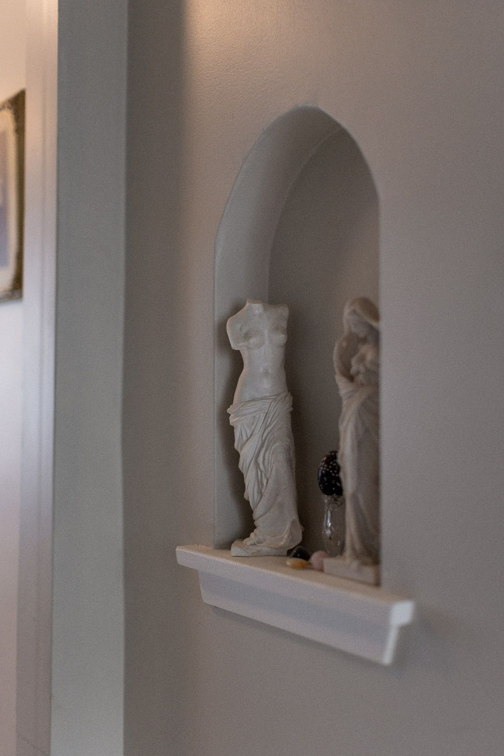 White classical Greek statues displayed in an arched wall niche with a small shelf in a minimalist interior.