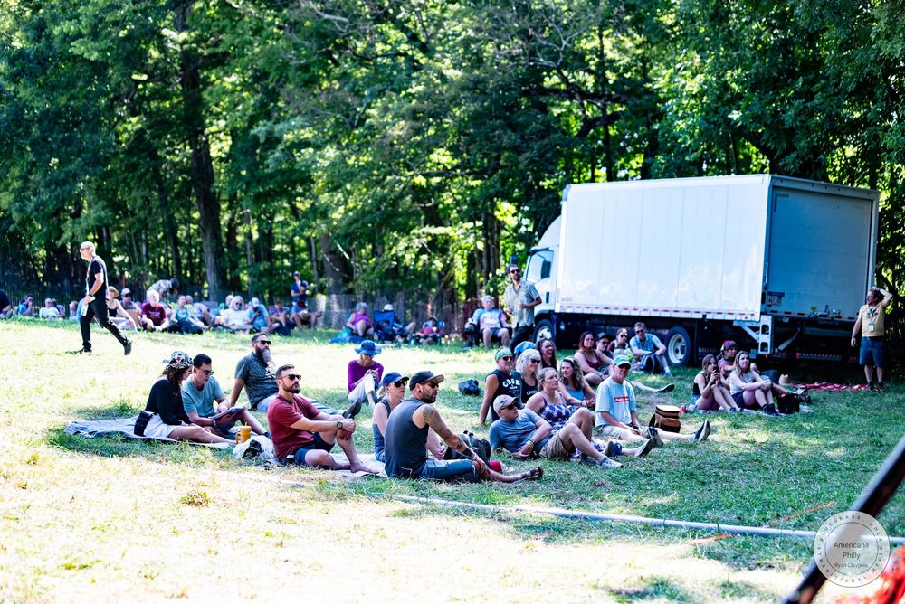 Crowd of people sitting on grass at an outdoor festival near a white truck surrounded by trees on a sunny day.