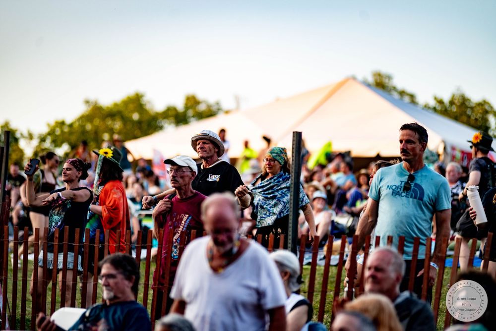 Crowd of festival-goers enjoying an outdoor music event near a white tent on a sunny evening.
