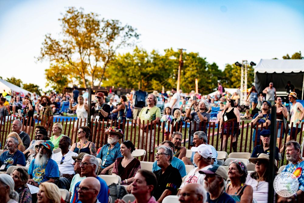 Large crowd enjoying an outdoor music festival on a sunny day with trees and a stage tent in the background.