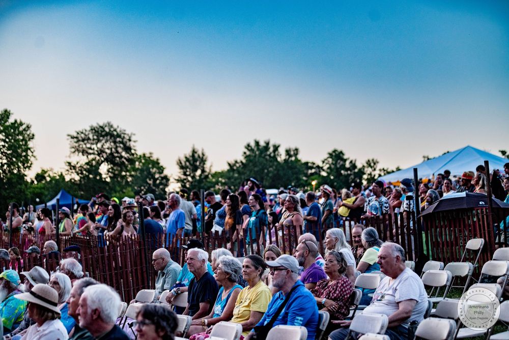 Large crowd of people seated and standing at an outdoor summer concert event at dusk with trees in background.