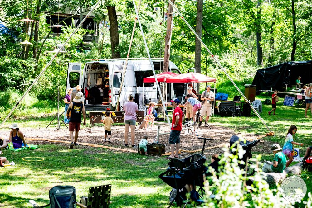 Families enjoying a sunny outdoor campground gathering with RVs, red canopy, and lush green trees.