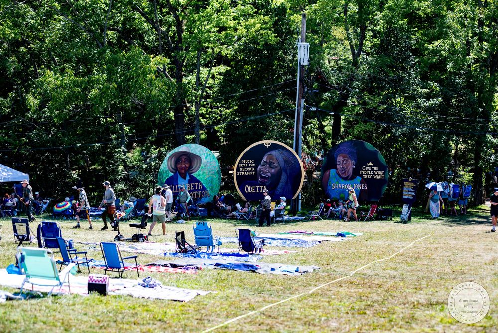 Outdoor jazz festival with large drum displays and lawn chairs on grass surrounded by lush green trees on a sunny day.