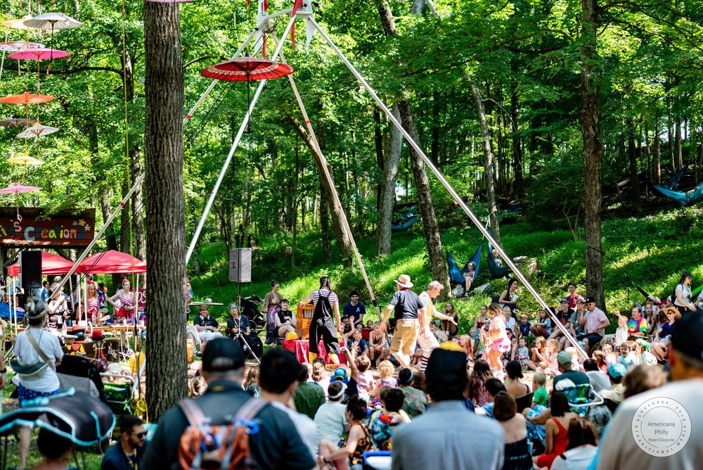 Outdoor festival crowd watches aerial performance on trapeze rig surrounded by lush green trees on a sunny day.
