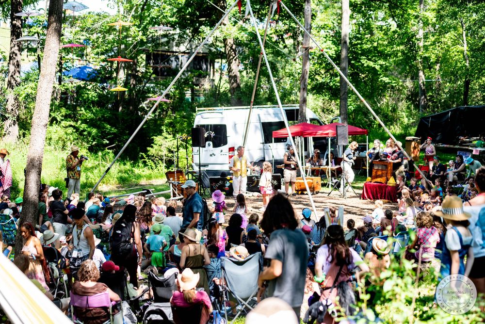 Outdoor music festival crowd gathers in a wooded area as a band performs on a rustic stage beneath a geometric structure.