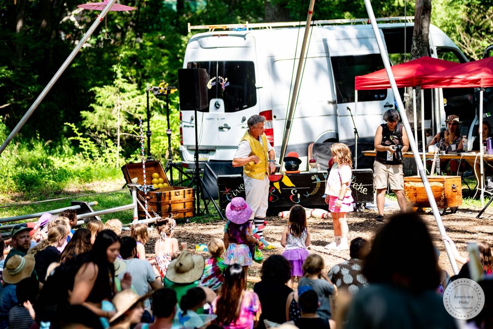 A clown entertains children at an outdoor festival with a camper and green trees in the background on a sunny day.