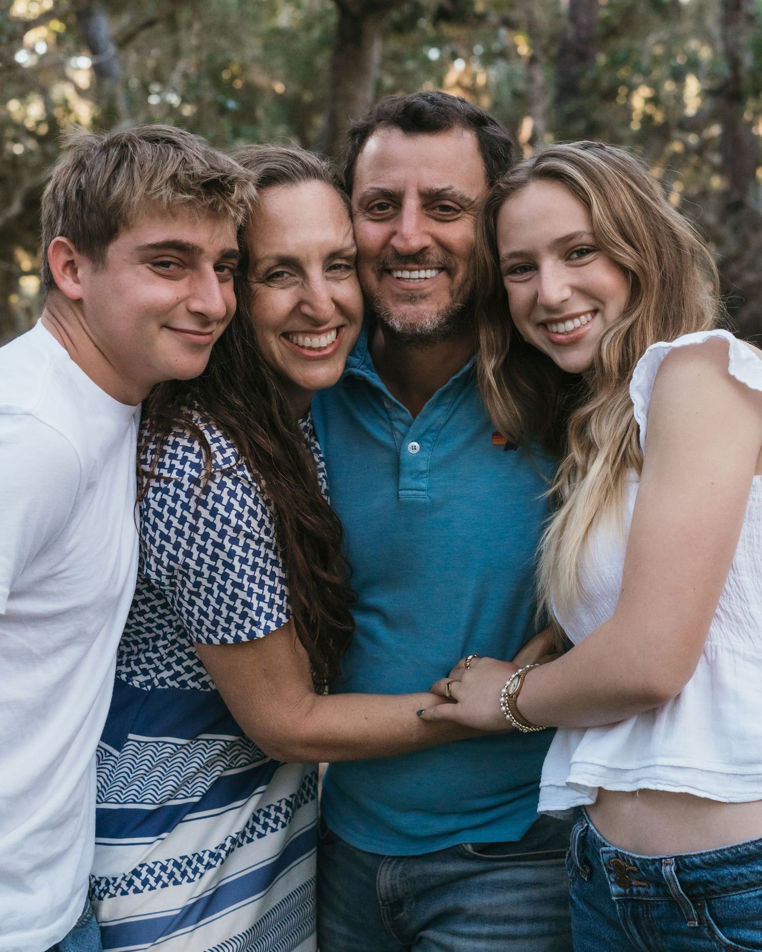 Happy family of four hugging and smiling outdoors among trees in a warm, candid portrait photo.