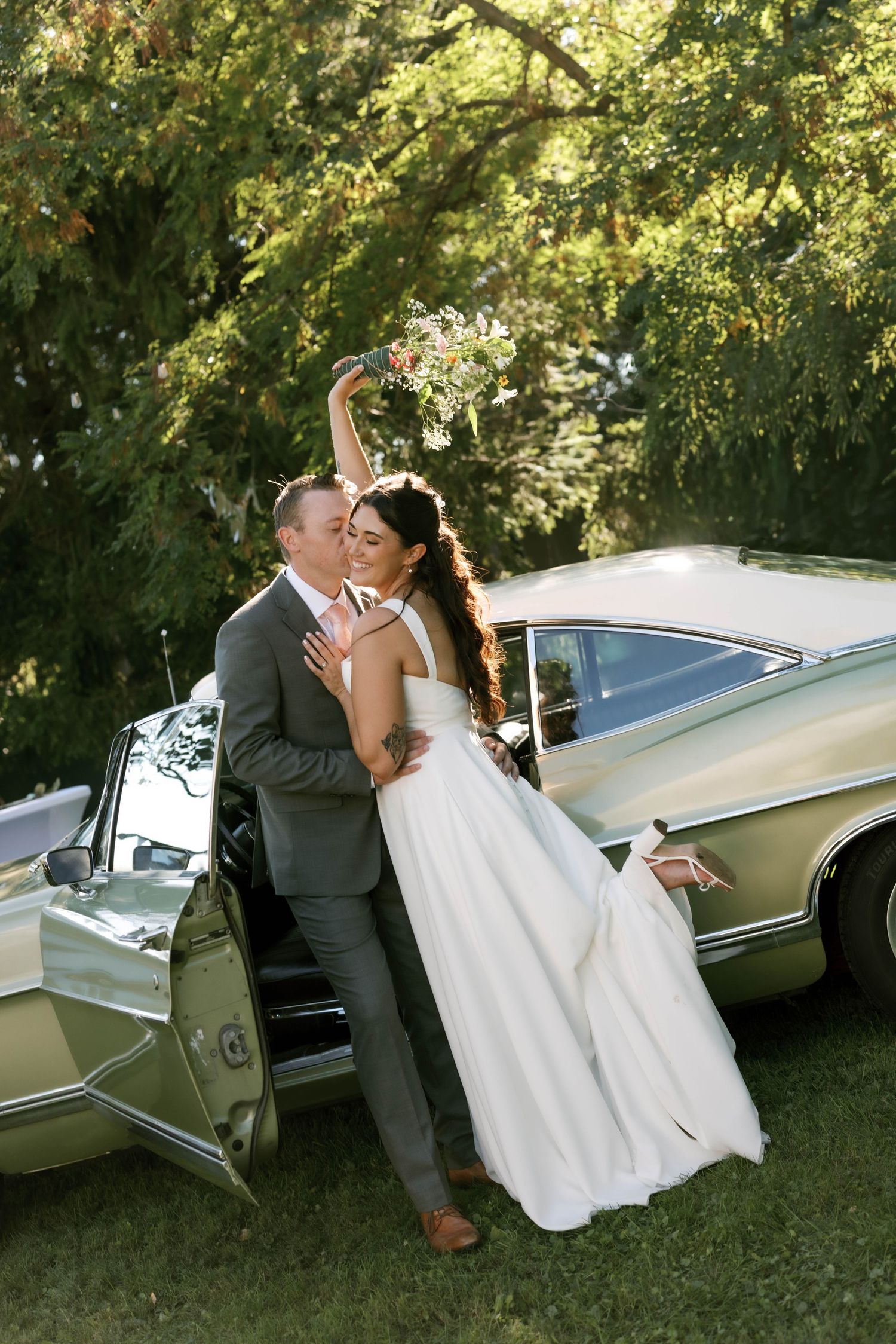 Bride and groom pose joyfully beside a vintage pale green car outdoors, surrounded by lush green trees on their wedding day.