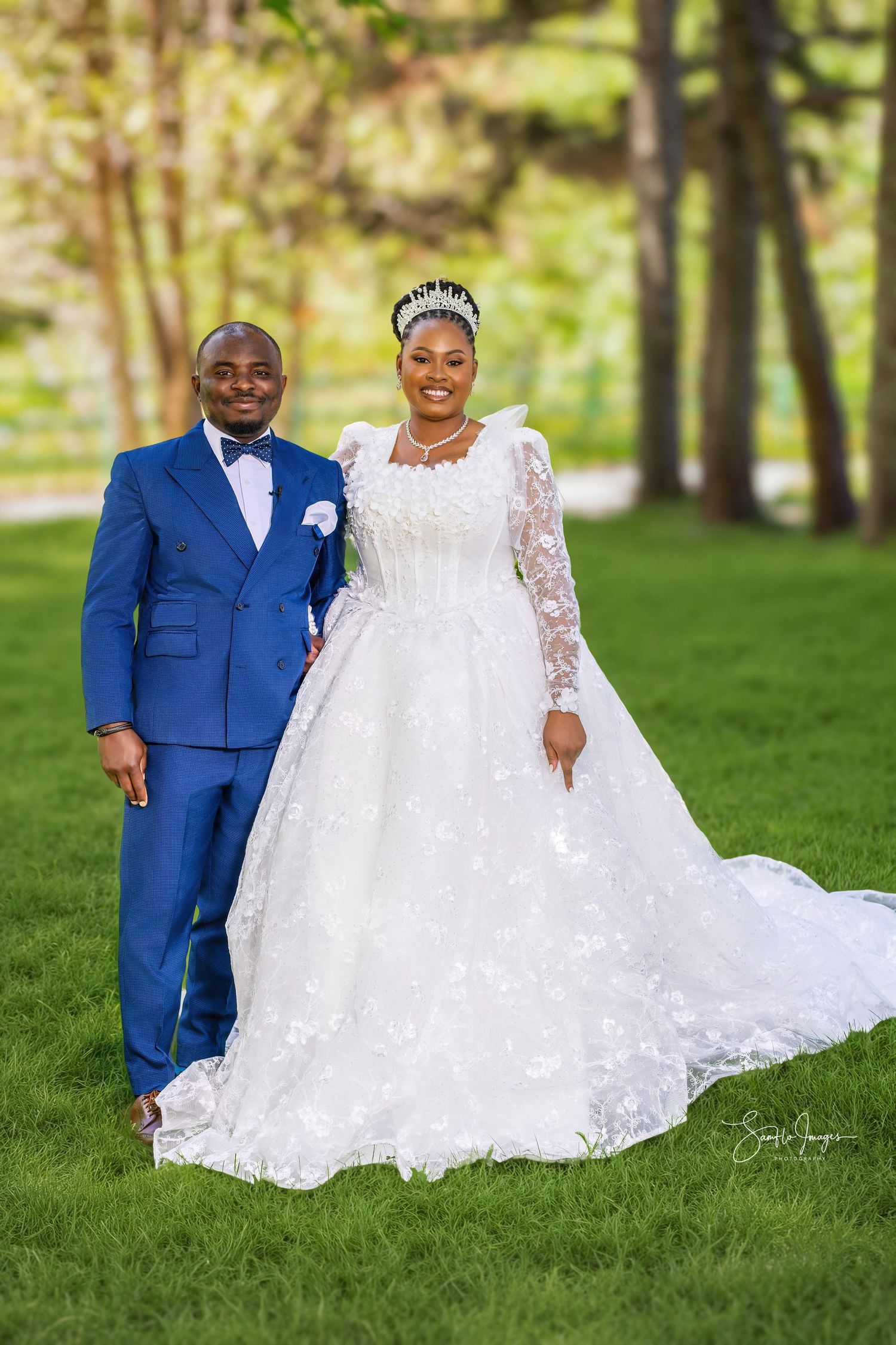 Elegant bride in white lace gown with tiara and groom in royal blue suit posing outdoors on green grass.