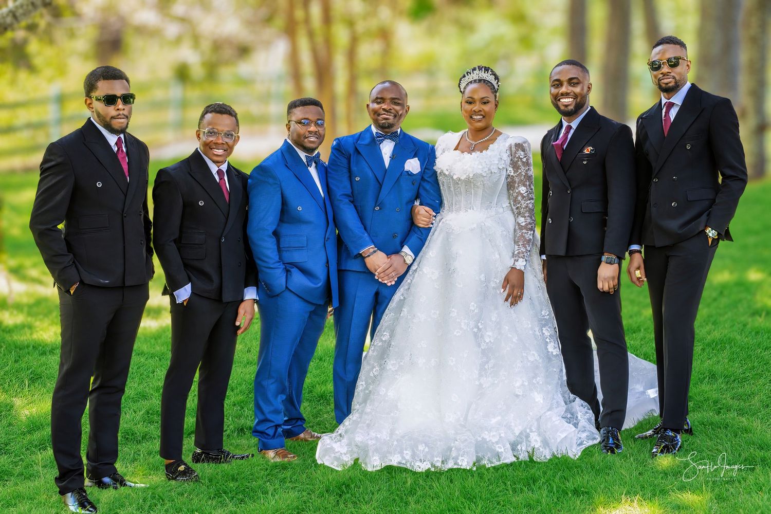 A bride in a white lace gown poses with groom in blue suit and groomsmen in black suits outdoors.