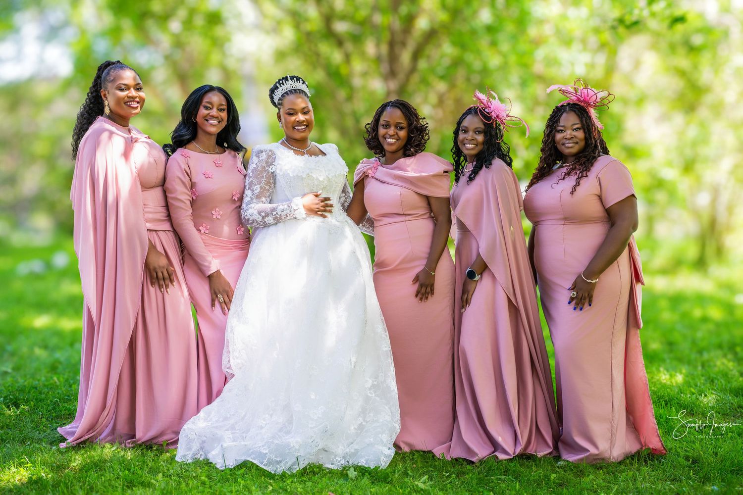Bride in white lace gown poses with five bridesmaids in pink dresses outdoors on a green lawn.