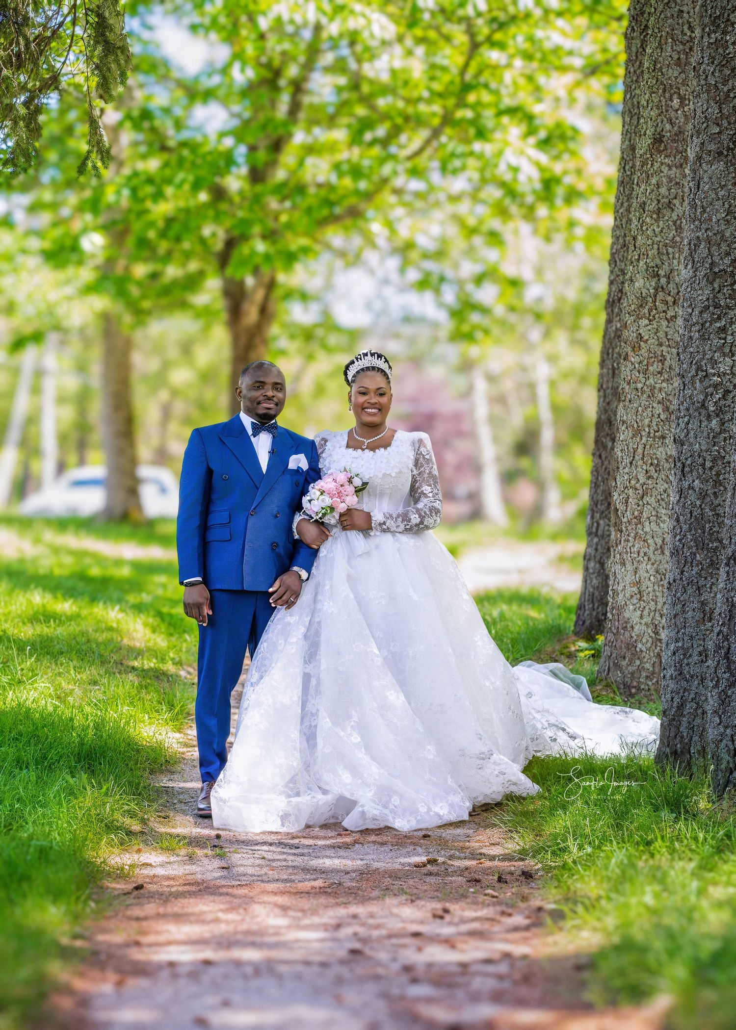 A couple poses on a tree-lined path, groom in blue suit and bride in white lace gown holding pink bouquet.