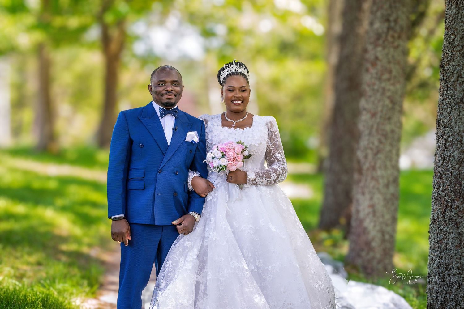 Bride and groom posing outdoors in park, bride in white lace gown holding pink bouquet, groom in blue suit.