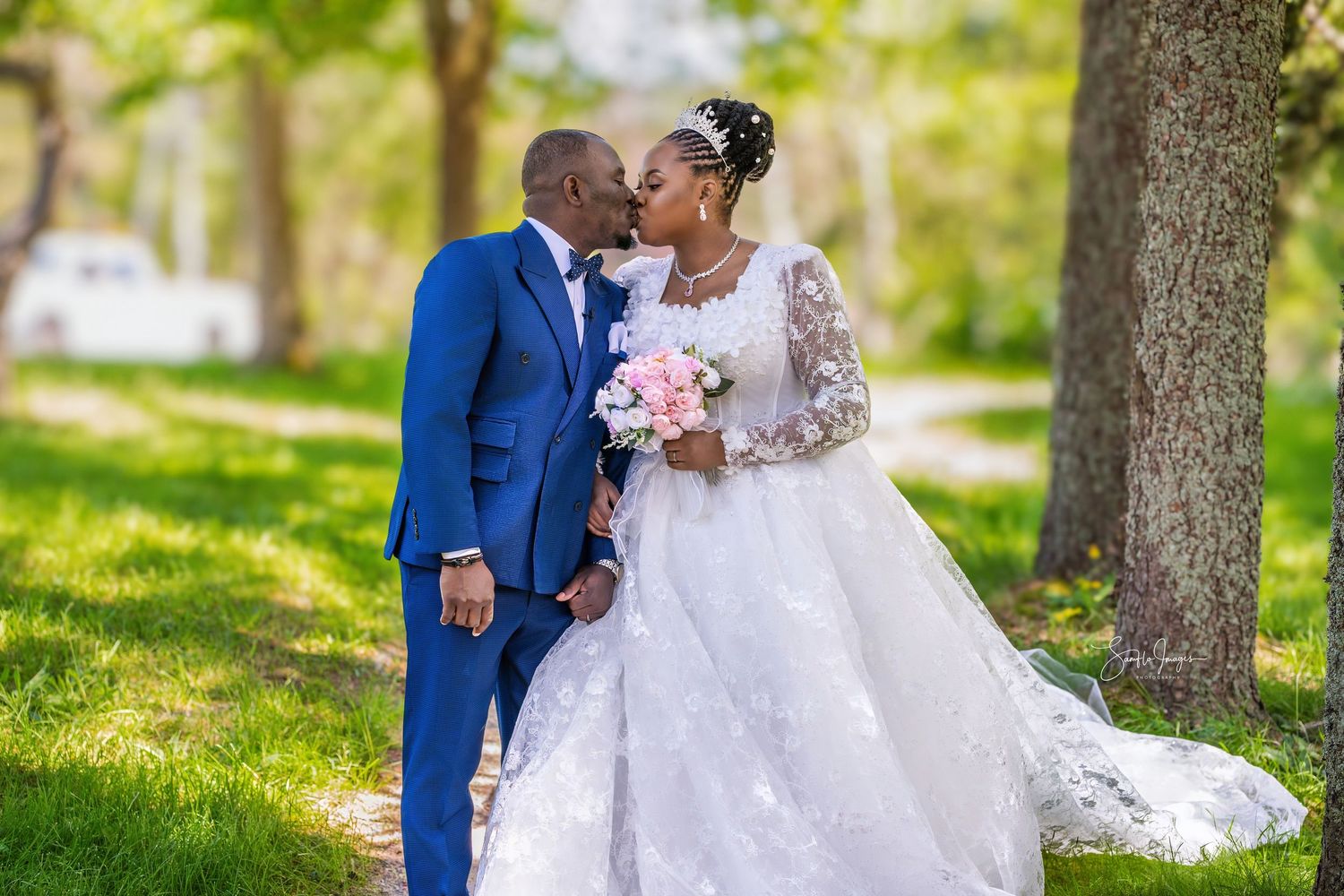 A bride and groom share a romantic kiss outdoors, surrounded by lush green trees, as she holds a pink bouquet.