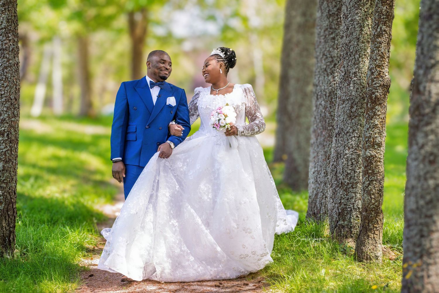 Bride and groom walking among trees in lush green park, wearing white lace gown and blue suit on wedding day.