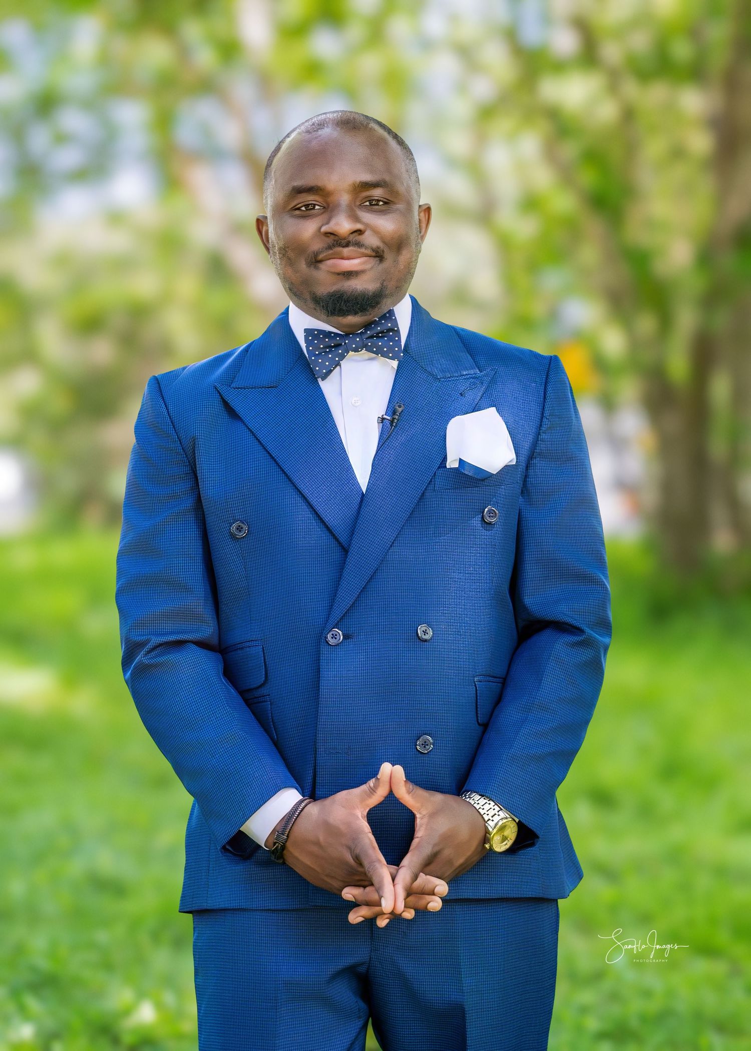Man in blue double-breasted suit with bow tie and white pocket square smiling outdoors in a green park setting.