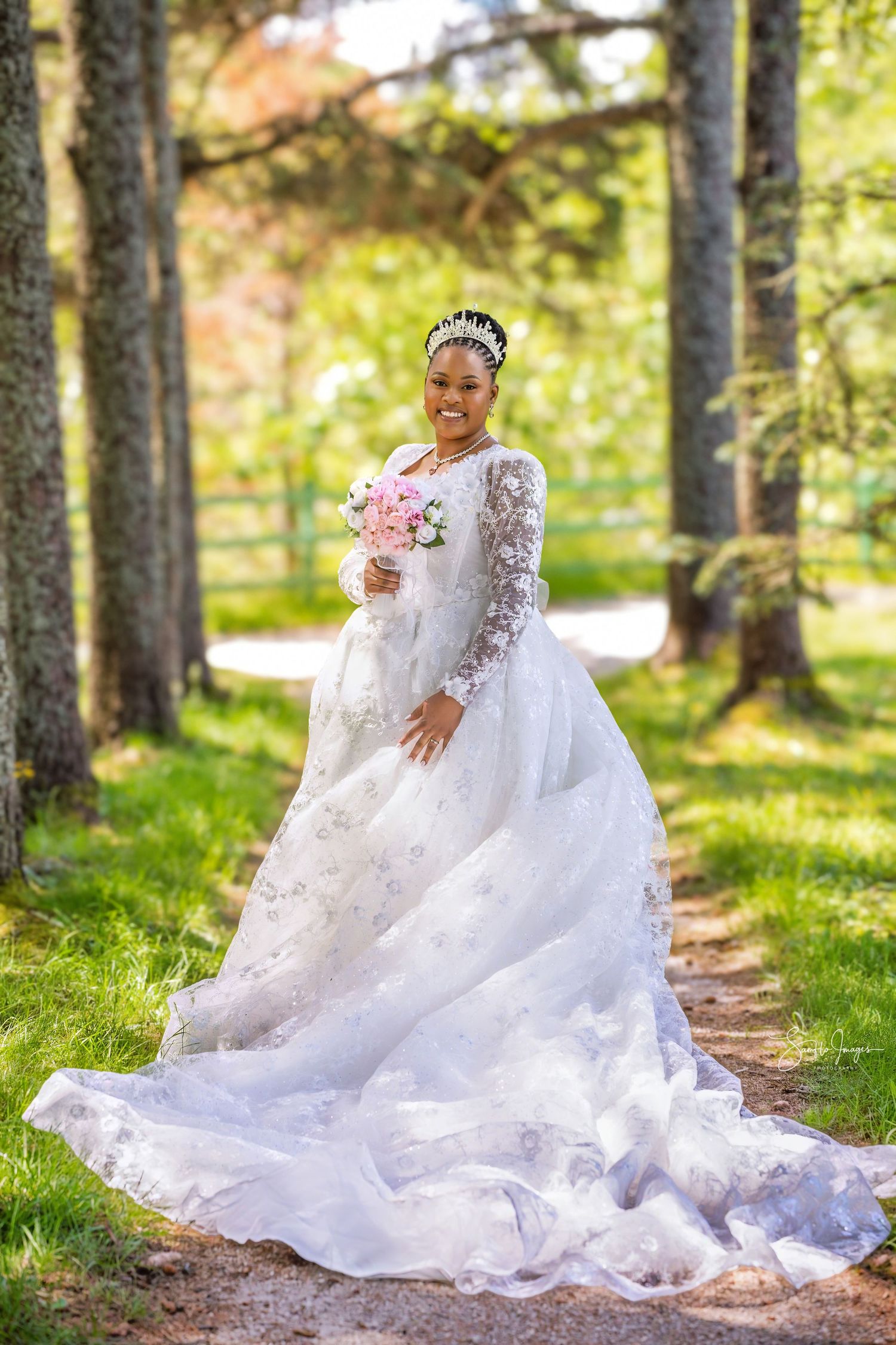Smiling bride in white lace gown with long train holds pink bouquet outdoors among tall trees on a sunny day.