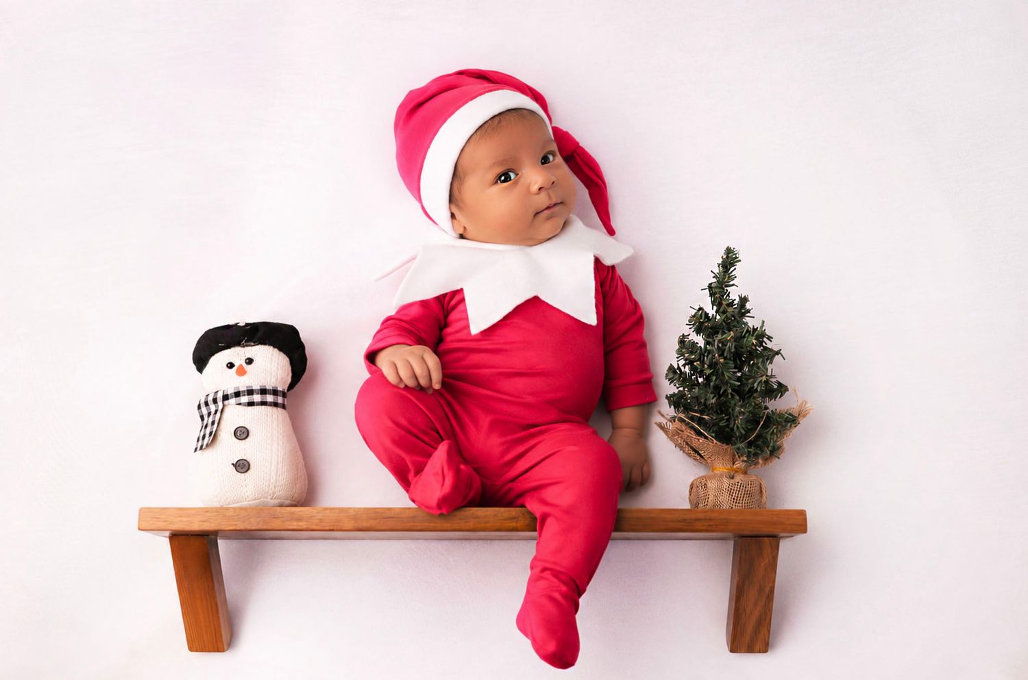 Baby in red elf outfit and Santa hat sits on wooden bench with snowman and mini Christmas tree.