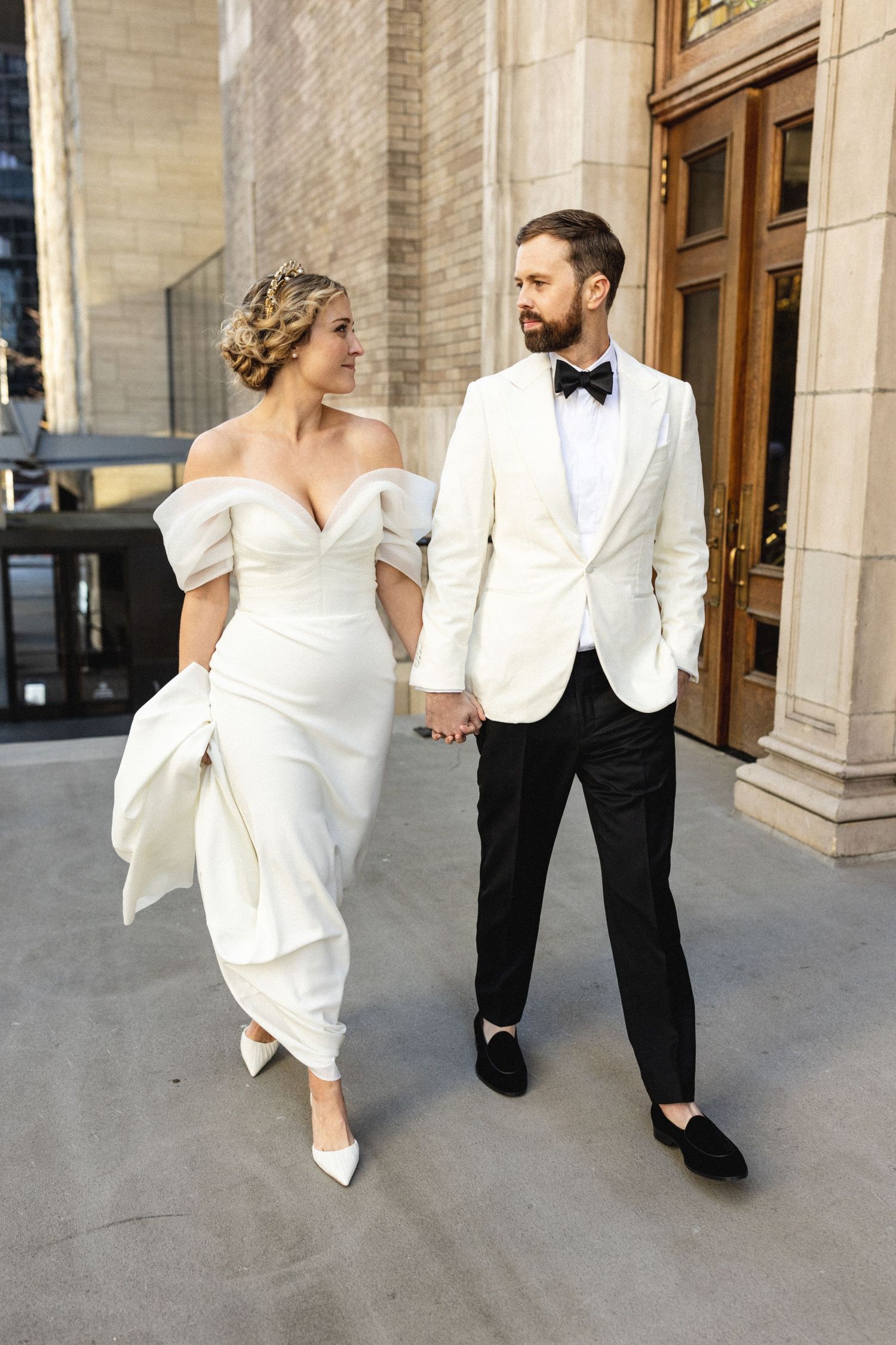 Bride in off-shoulder white gown holds hands with groom in white tuxedo jacket walking near elegant building.