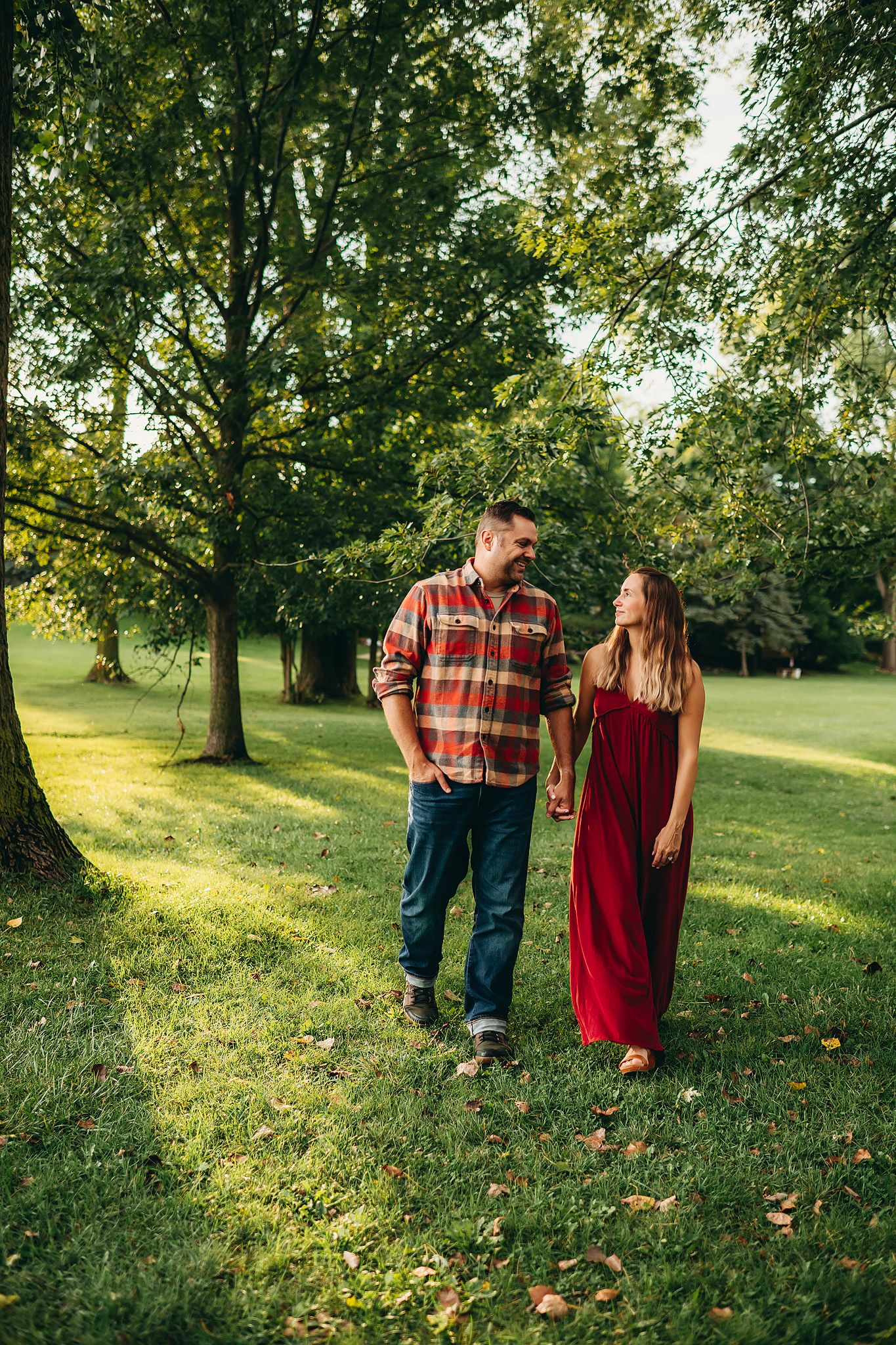 Couple holding hands and smiling while walking through a lush green park, woman in red dress and man in plaid shirt.