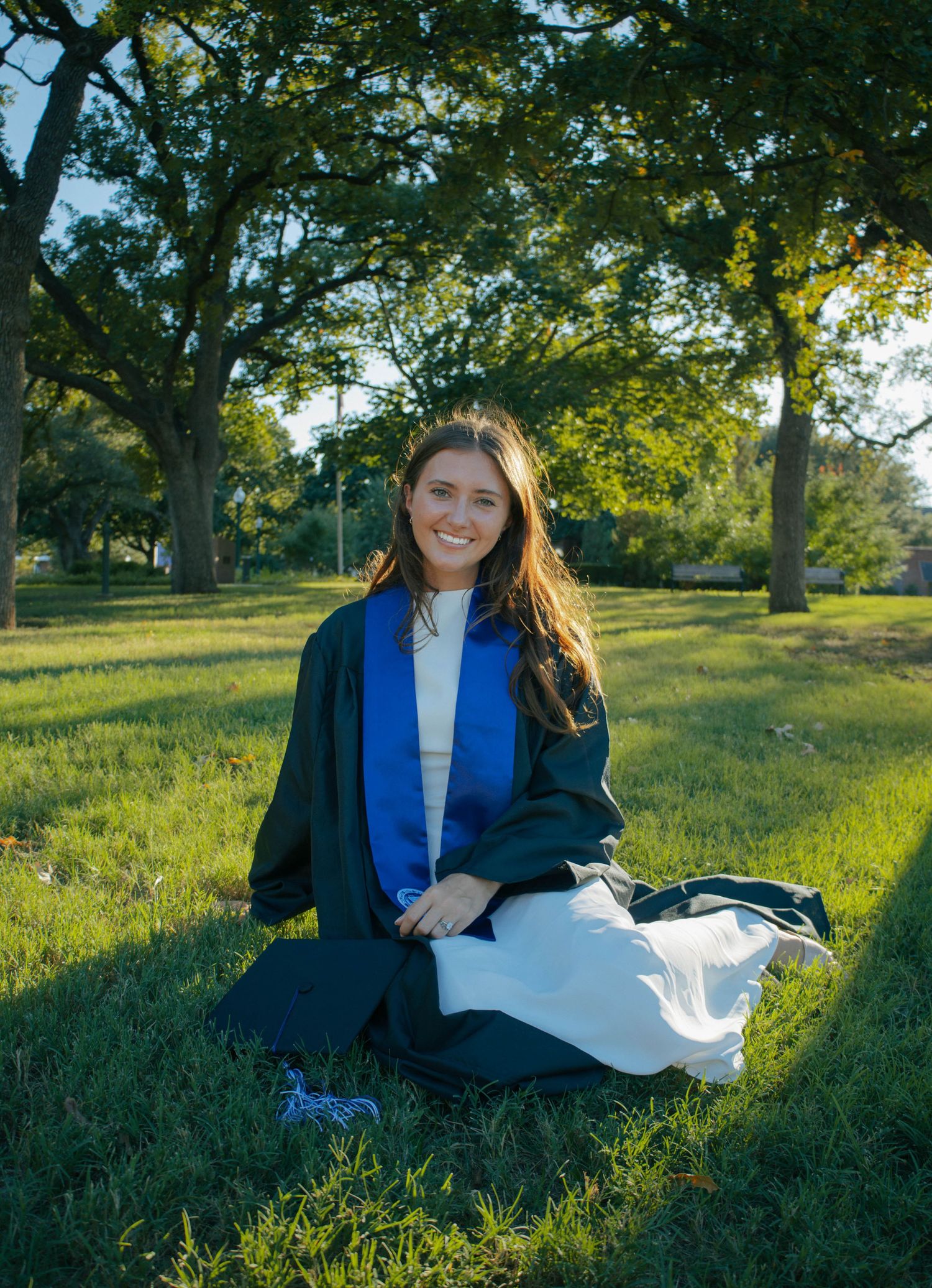 Smiling graduate in green cap and gown sitting on lush grass outdoors with trees in background on sunny day.
