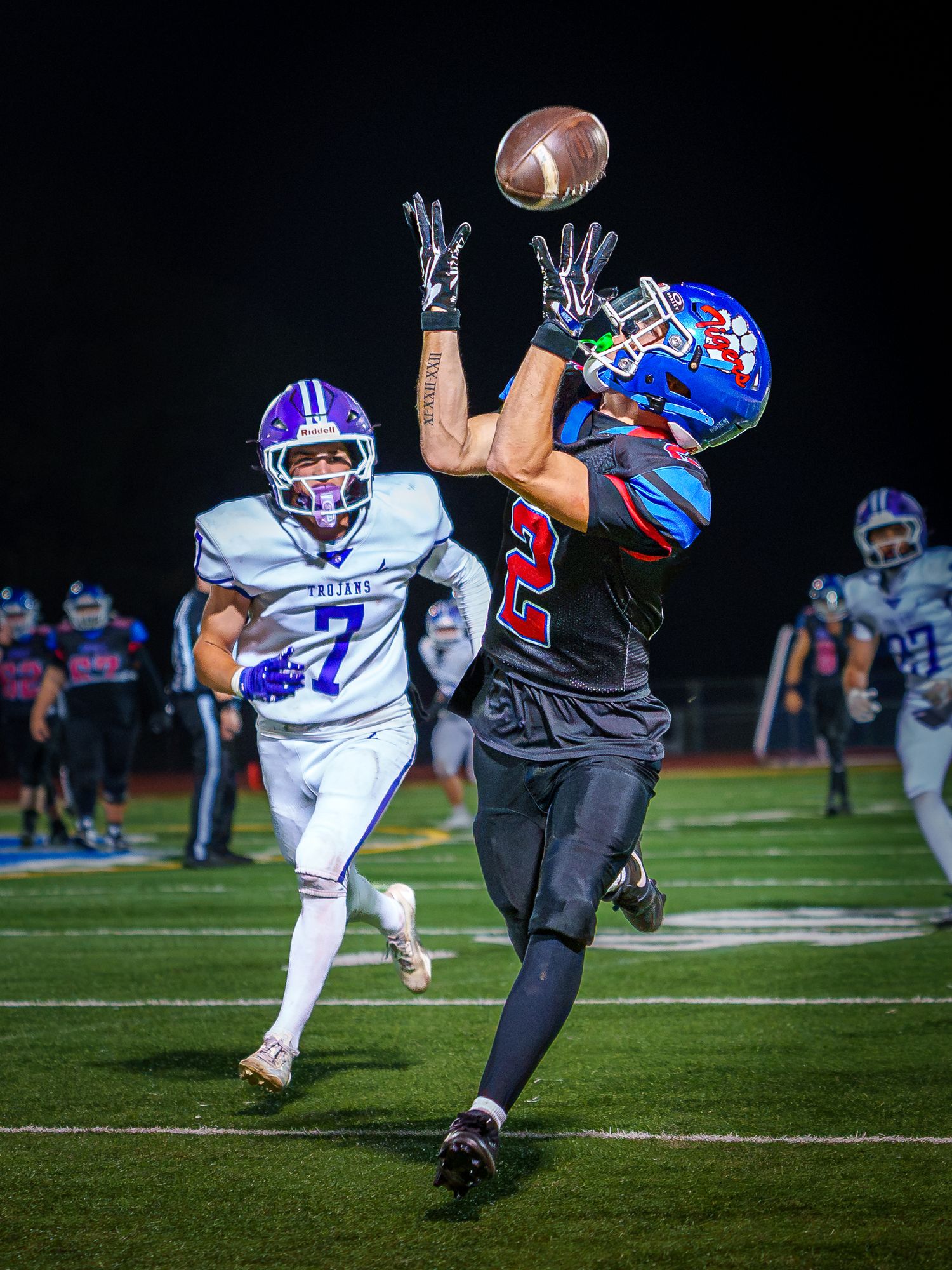 Football player in red and blue uniform catches a pass while defender in purple jersey pursues during night game.