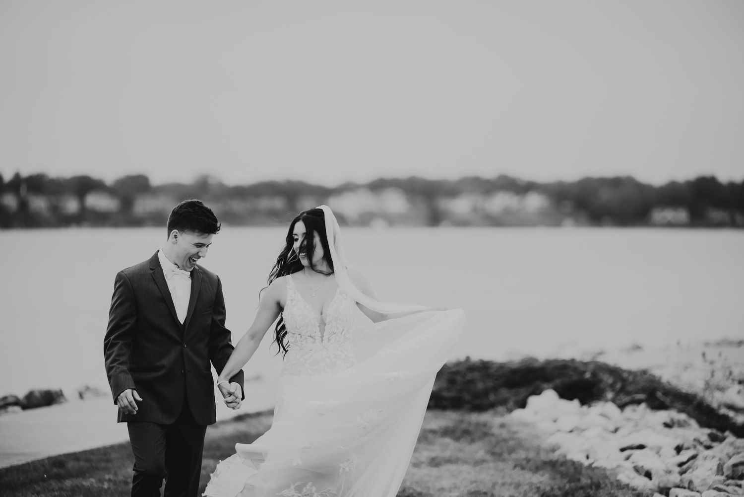 Black and white photo of a bride and groom walking hand in hand by a lake, her gown flowing in the wind.