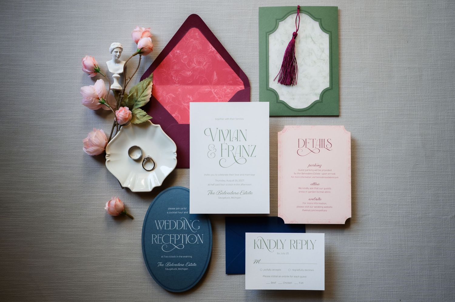 Elegant wedding invitation suite with navy, green, and pink stationery, gold rings, and floral accents on a gray background.