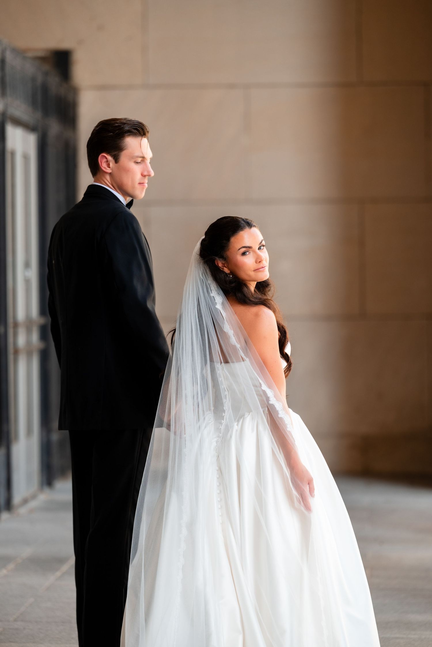 Bride in white ball gown and veil stands with groom in black tuxedo during elegant wedding portrait session.