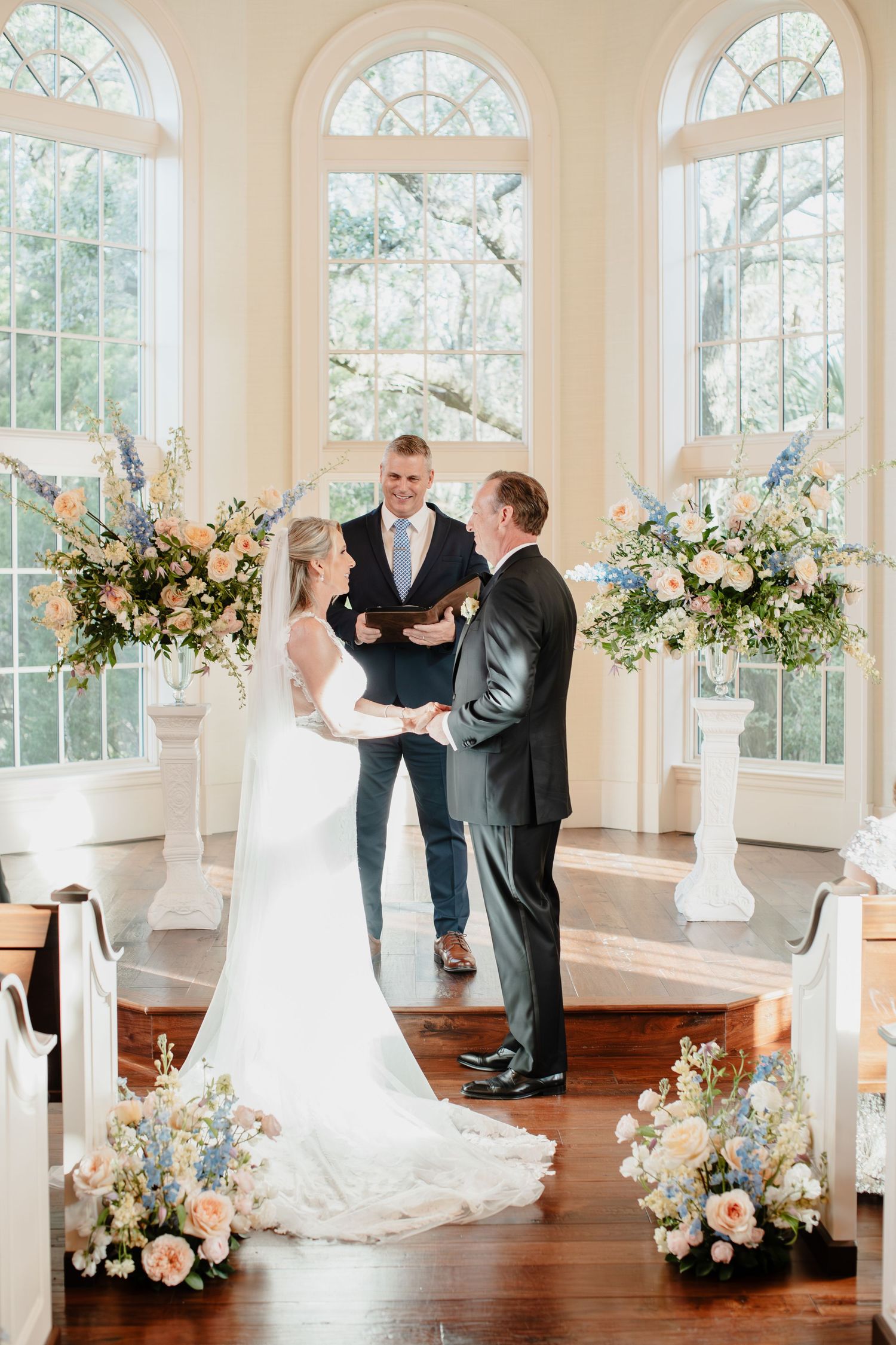 Bride and groom exchange vows at elegant chapel altar adorned with pastel floral arrangements of pink, blue, and white blooms.