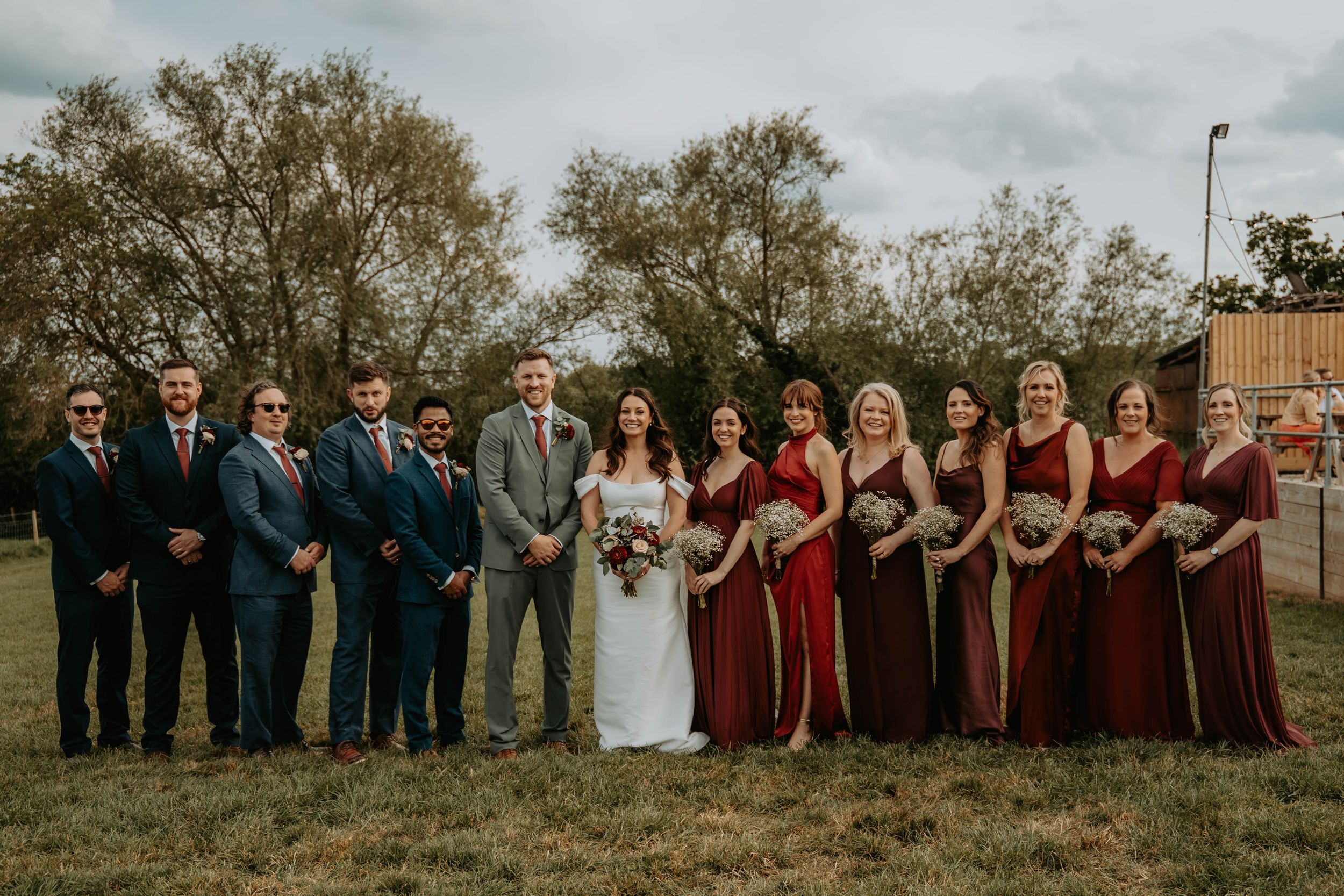 Wedding party posing outdoors in burgundy and navy attire, bride in white gown holding bouquet, surrounded by groomsmen and bridesmaids.