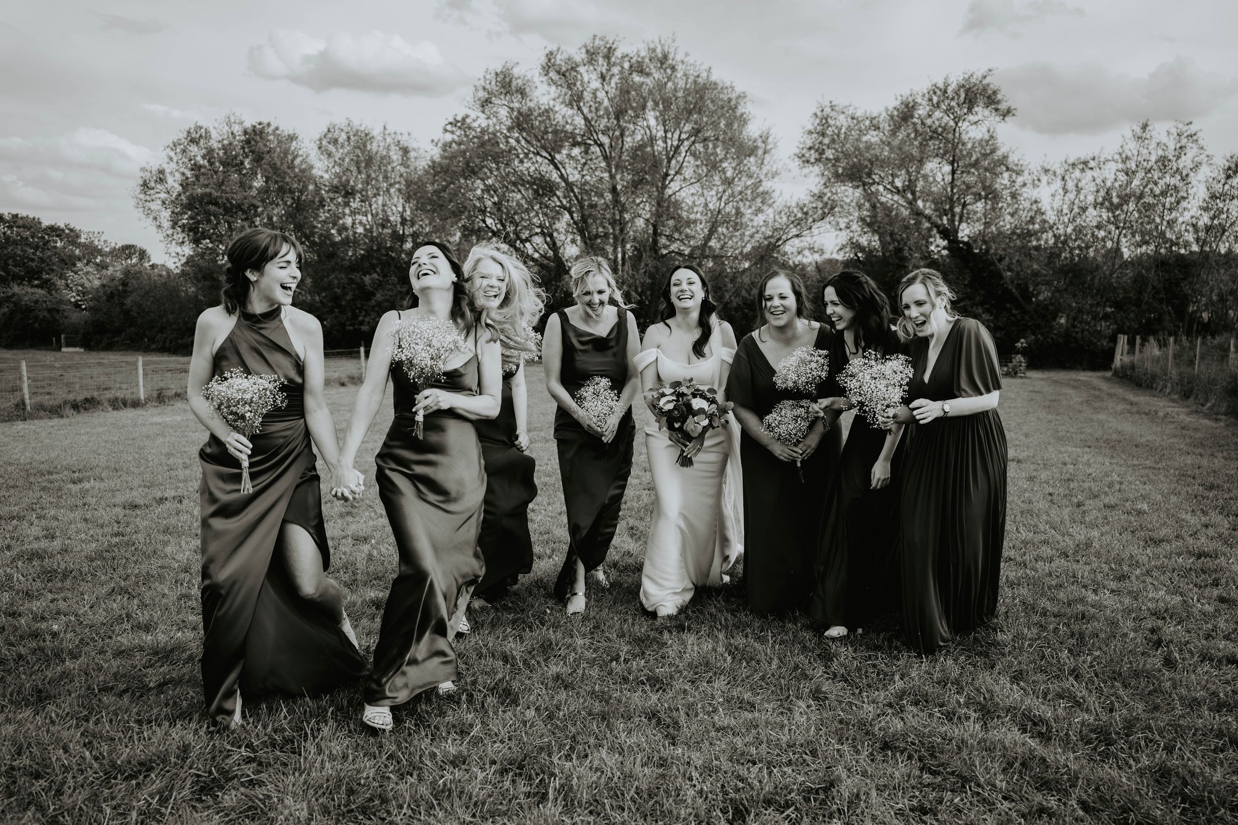 Black and white photo of a bride and bridesmaids laughing and walking together outdoors at a wedding.