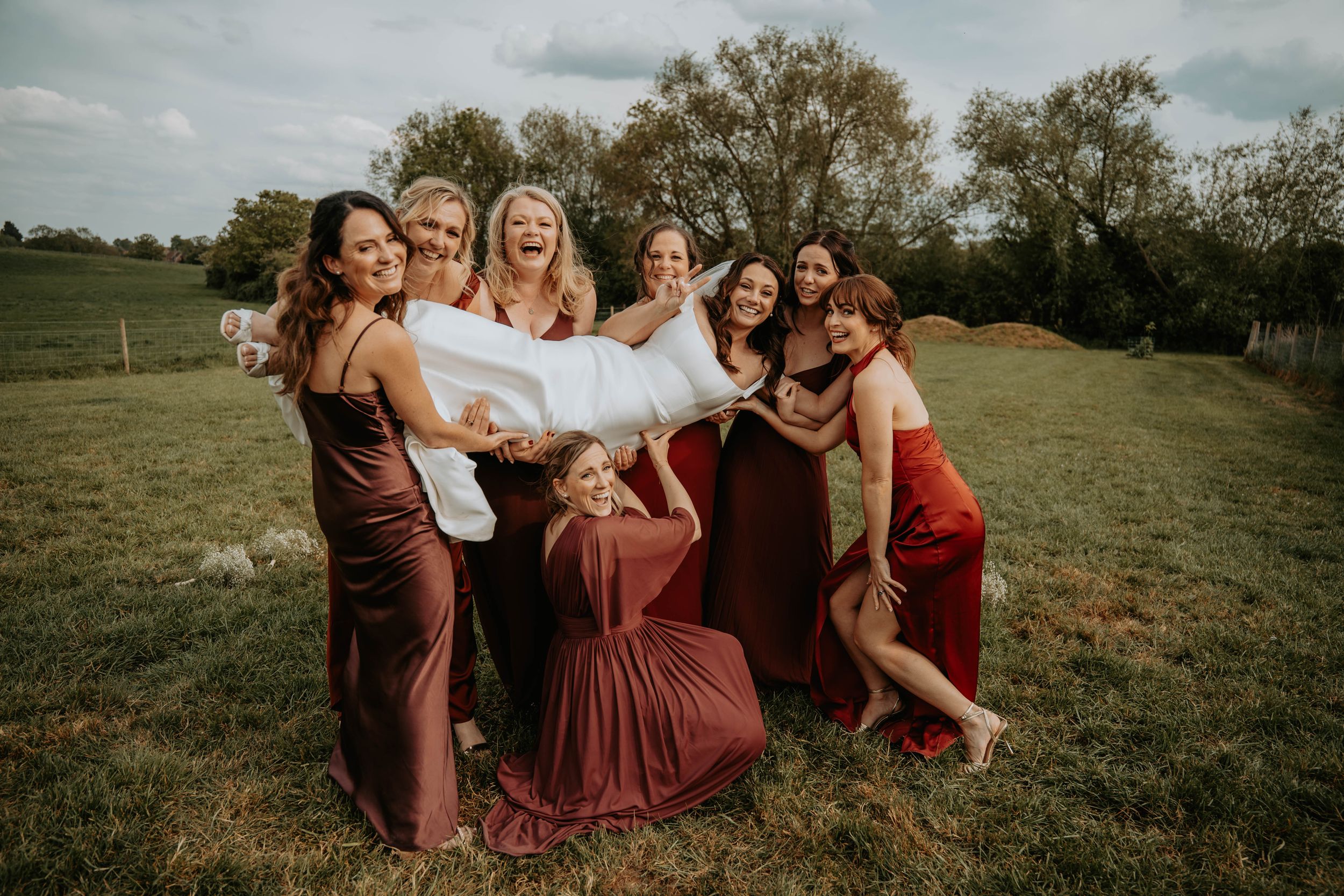 Bridesmaids in burgundy and red dresses joyfully lifting bride in white gown on green field outdoors.
