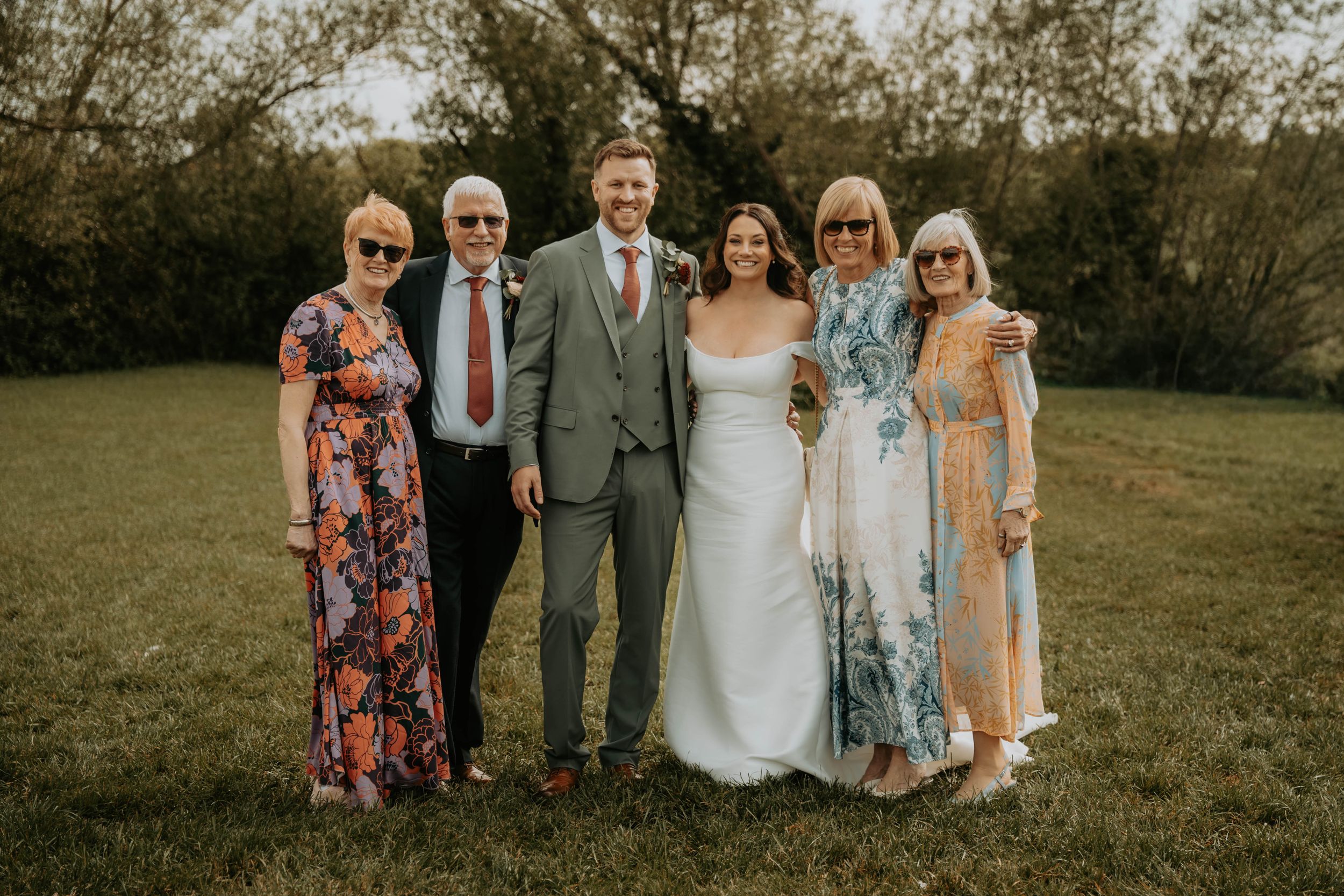A bride and groom pose with three family members on a green lawn at an outdoor wedding celebration.