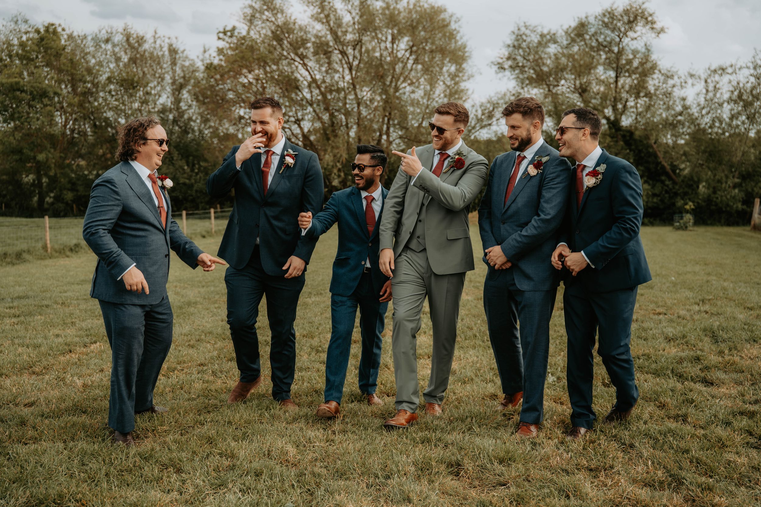 Groomsmen in navy and gray suits laughing together outdoors on a green field at a wedding celebration.