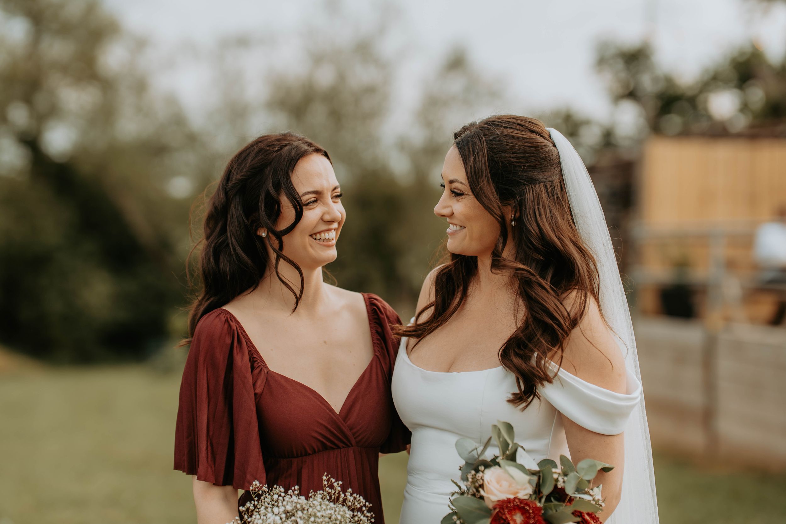 Bride in white gown with veil holding bouquet smiles at bridesmaid in burgundy dress outdoors.