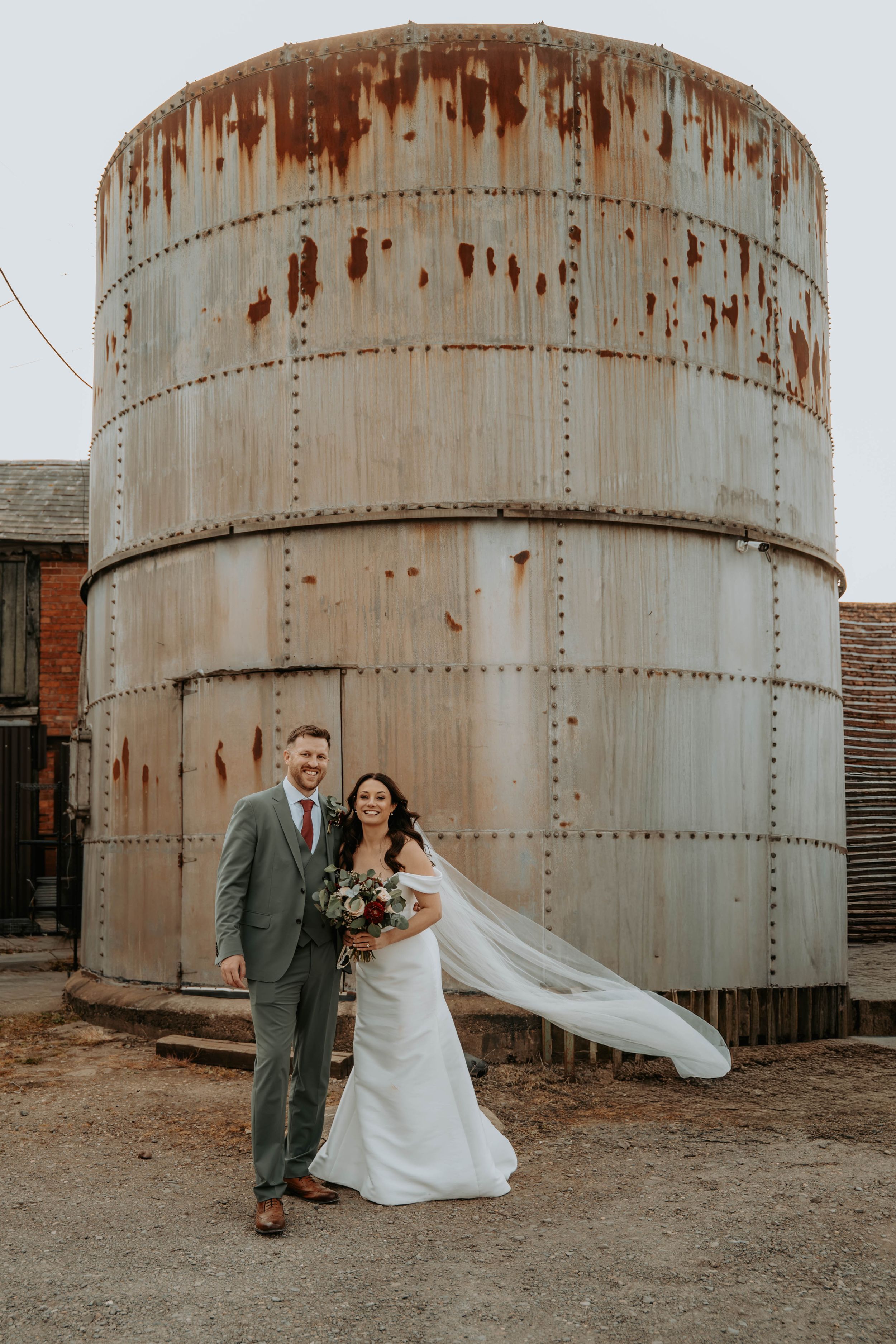 Bride and groom pose in front of a rustic metal silo at an industrial wedding venue, veil flowing in the wind.