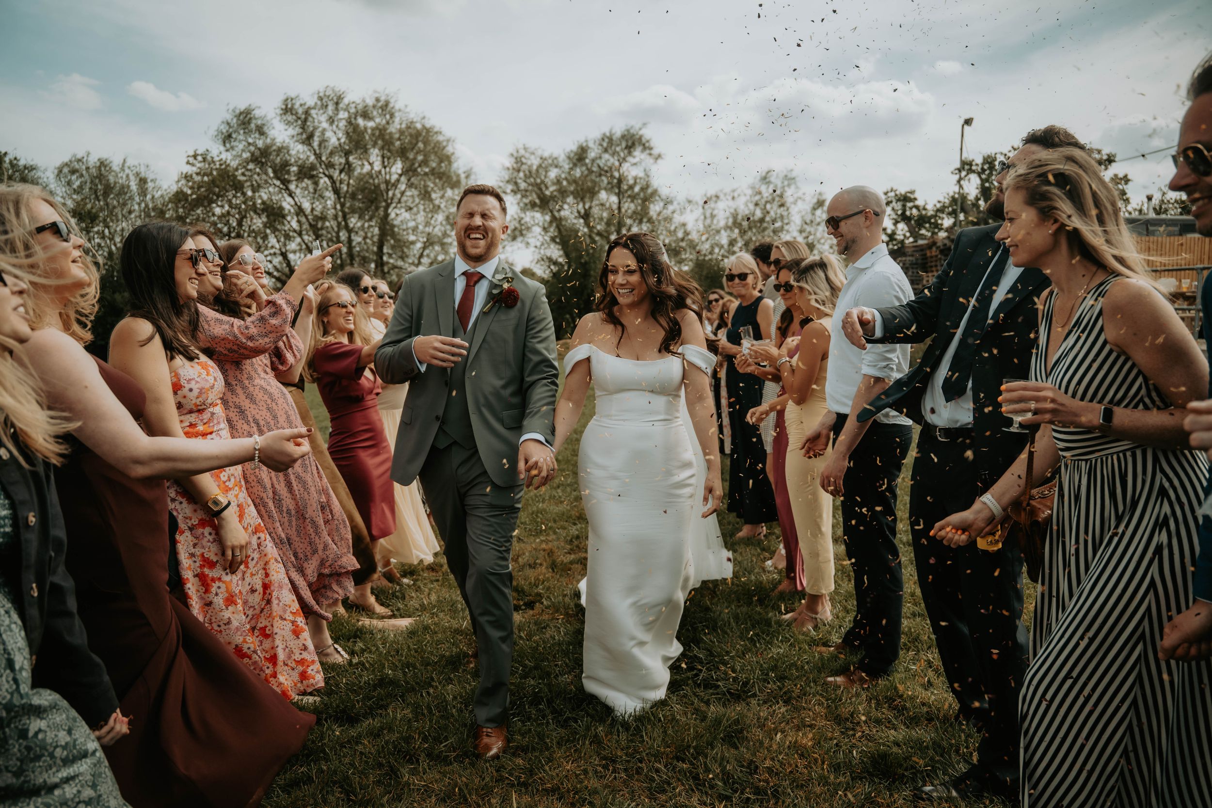 Bride and groom walk through confetti tunnel surrounded by cheering wedding guests outdoors.