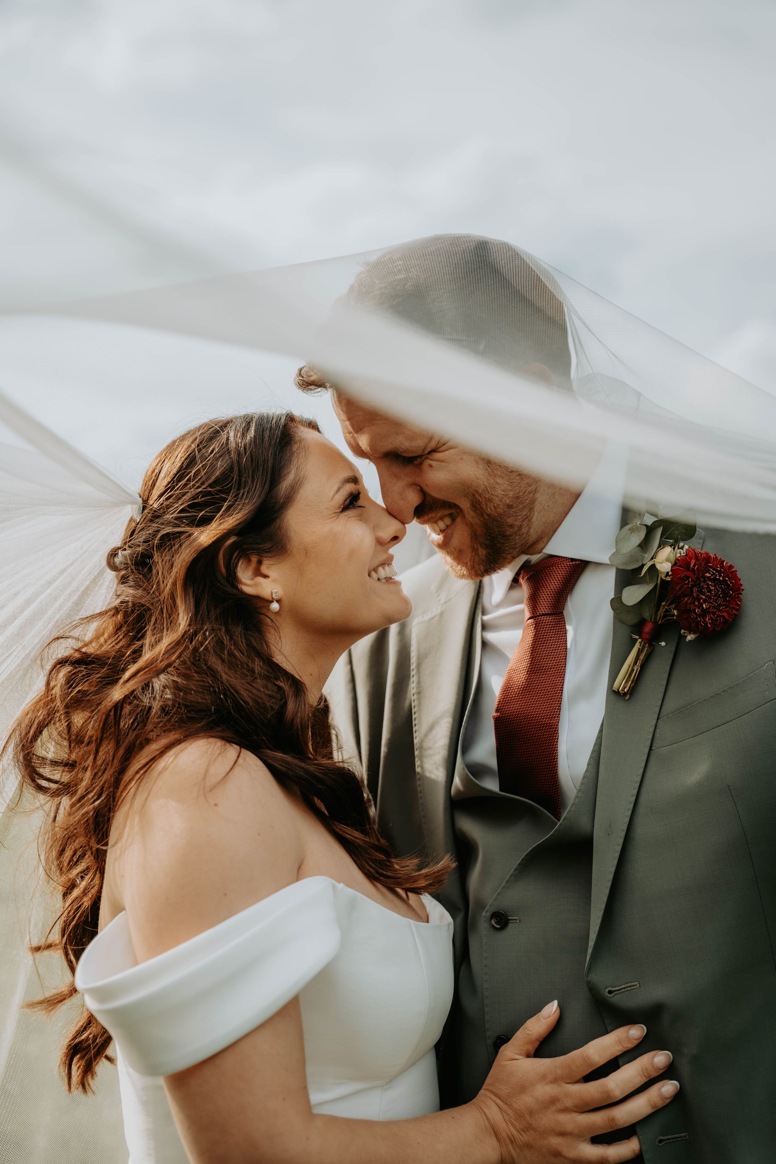 Bride and groom smiling face-to-face under flowing veil, groom in gray suit with red tie and boutonniere.