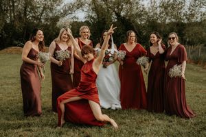 Bride in white gown poses with bridesmaids in burgundy and mauve dresses holding bouquets outdoors.