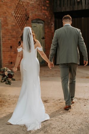 Bride and groom walking hand in hand away from camera, bride in white gown with veil holding bouquet, groom in grey suit.