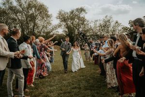 Bride and groom walk through a joyful confetti send-off surrounded by smiling guests on a lush green lawn.