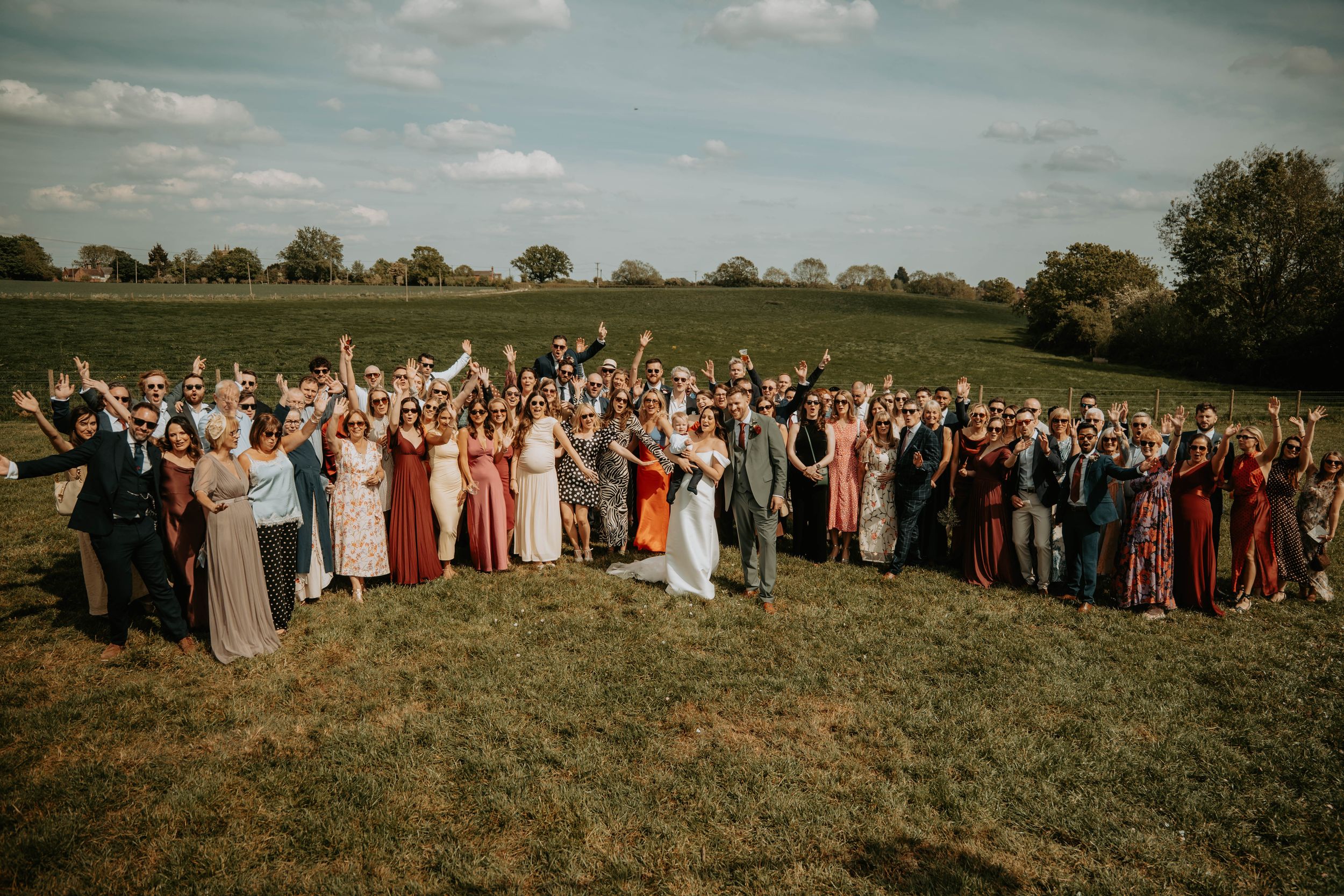 Large wedding party celebrating outdoors in a green field, guests in colorful dresses raising hands joyfully.