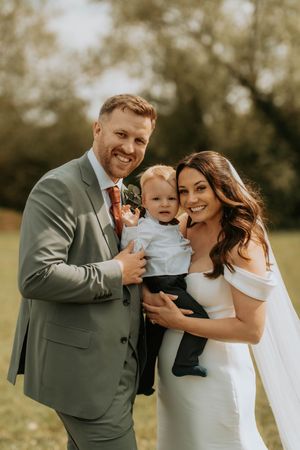 Smiling bride and groom in wedding attire holding their baby boy outdoors on their wedding day.