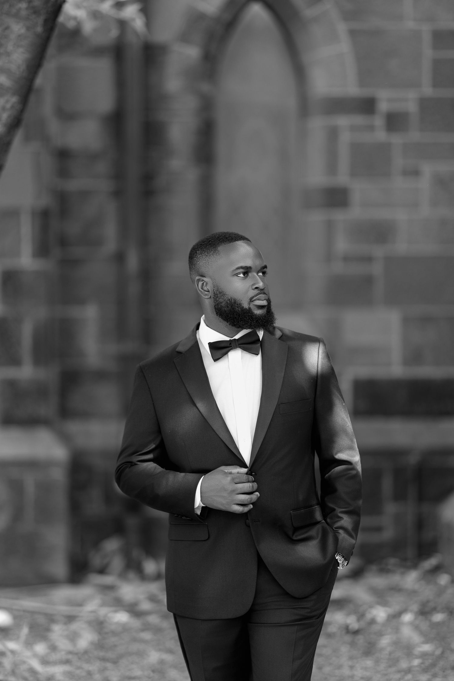 A well-dressed Black man in a black tuxedo with bow tie stands confidently outside a stone church building.