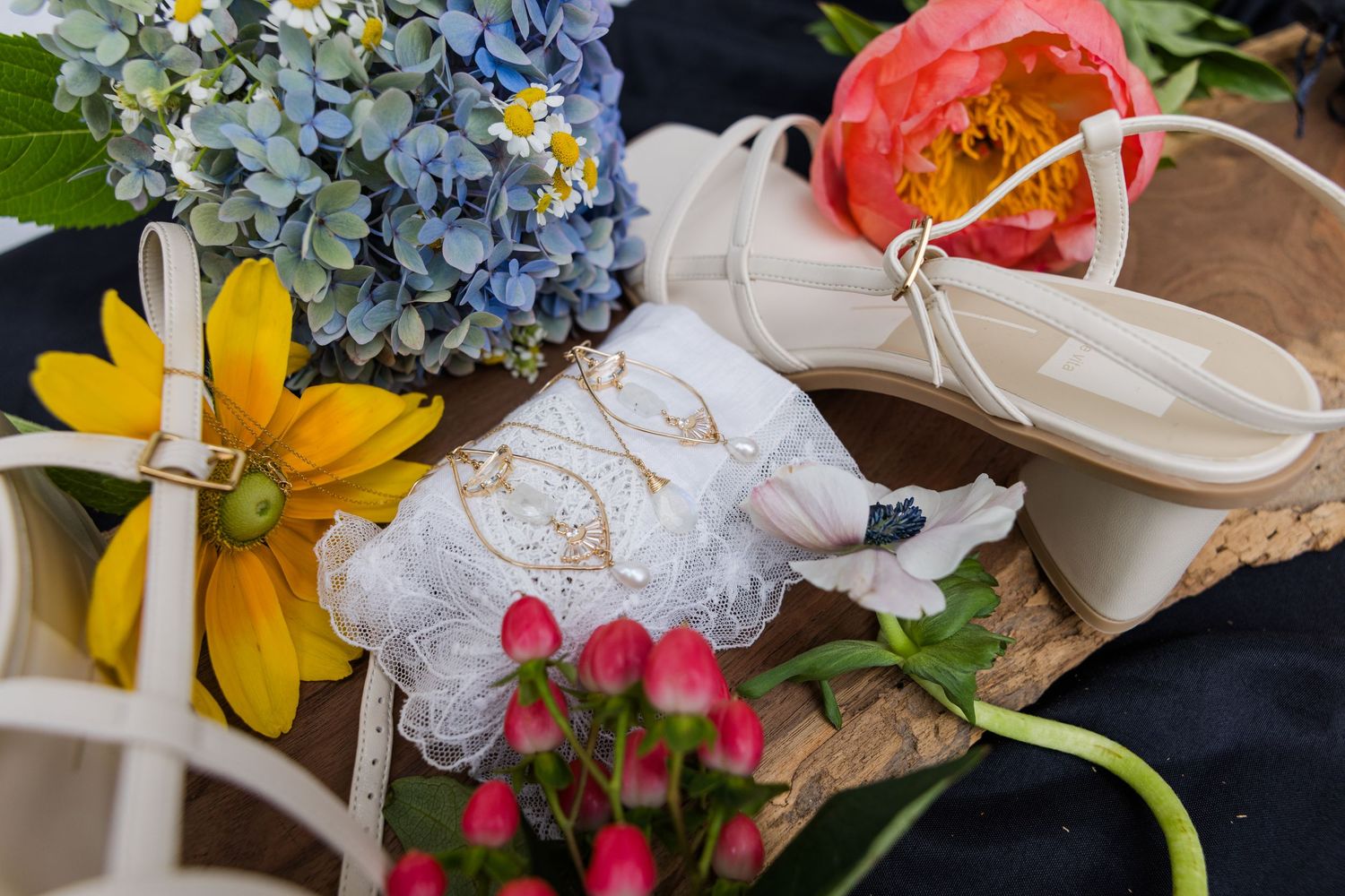 Bridal flat lay featuring embroidered garter, strappy heels, and colorful flowers including hydrangeas and peonies.