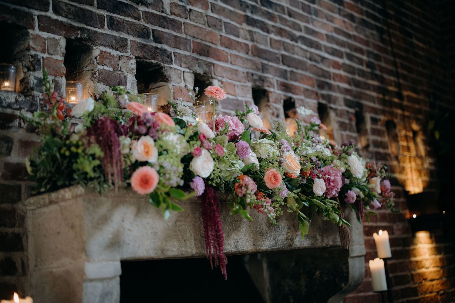 Lush floral arrangement of pink, white, and purple blooms cascading over a rustic brick fireplace mantel with candles.